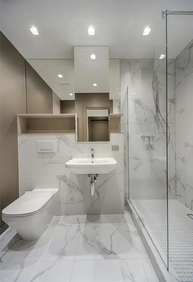 Baño pequeño en mármol blanco con inodoro, lavabo suspendido y ducha con mampara de cristal